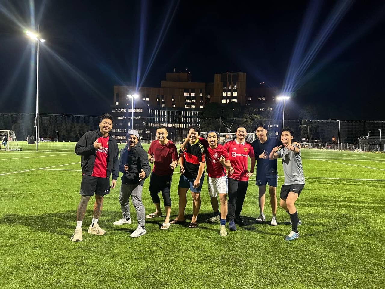 Seven players grinning on the pitch under floodlight beams