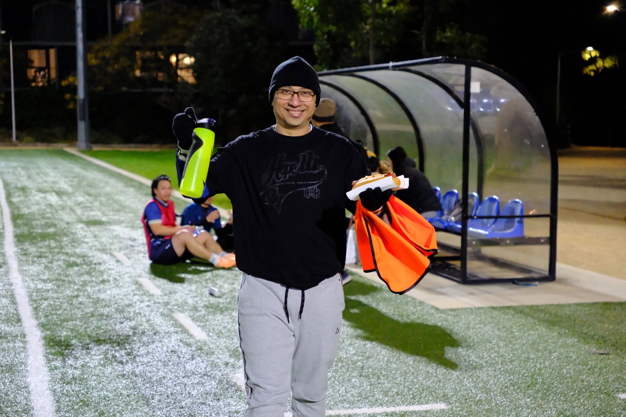 Player in black holding bibs and a water bottle beside the dugout