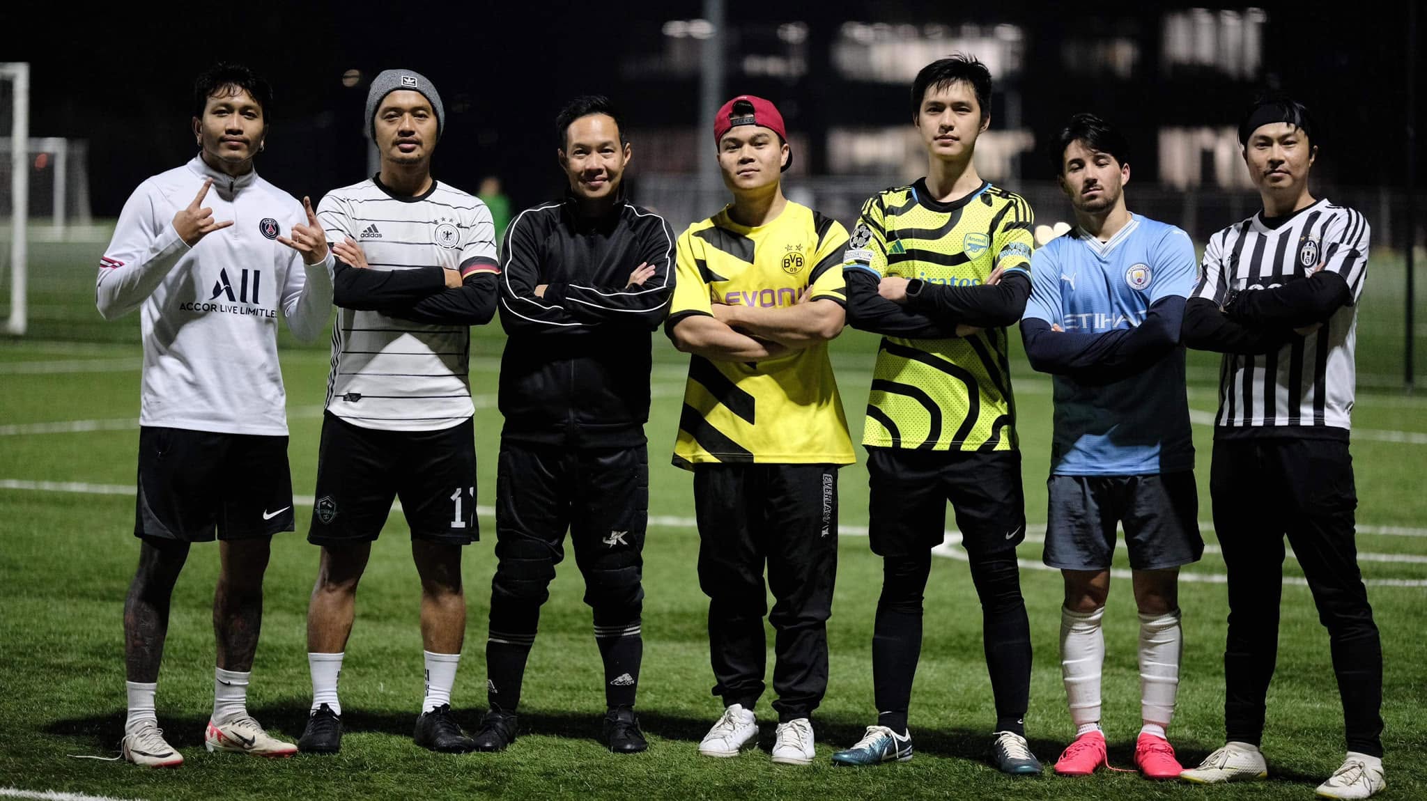 Seven MCFC players in club and favourite-team shirts, arms crossed on the night pitch